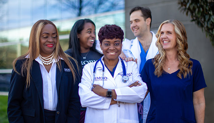 A group of health care professionals stand in front of Emory Hillandale Hospital in a group smiling 