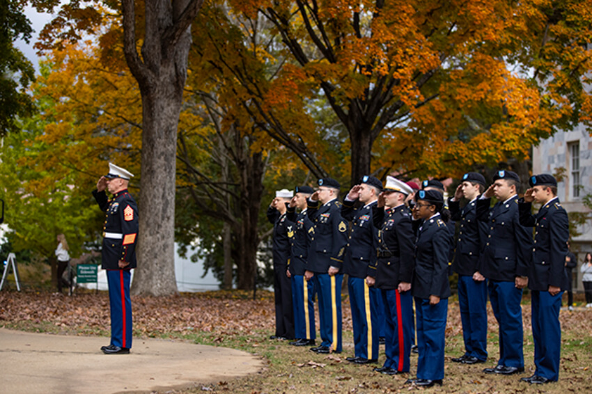 Veterans in uniform stand in rows and salute under fall leaves